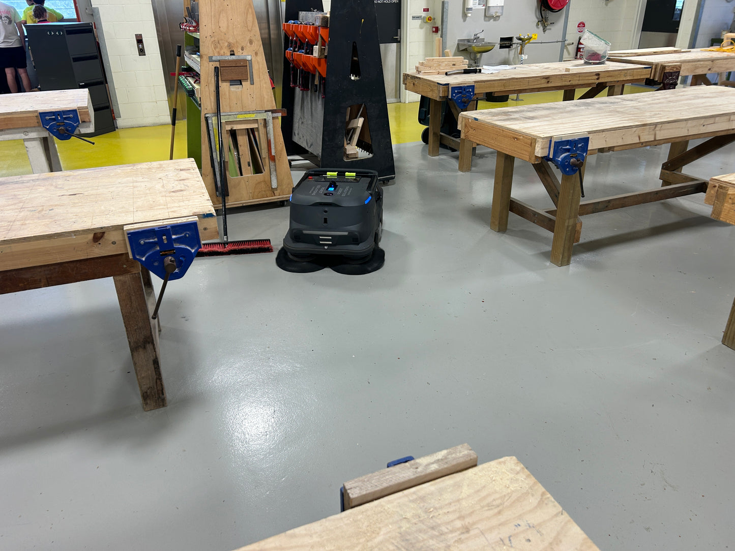 Robotic vacuum cleaner on a school workshop floor with wooden workbenches and tools. From Robot Solutions QLD